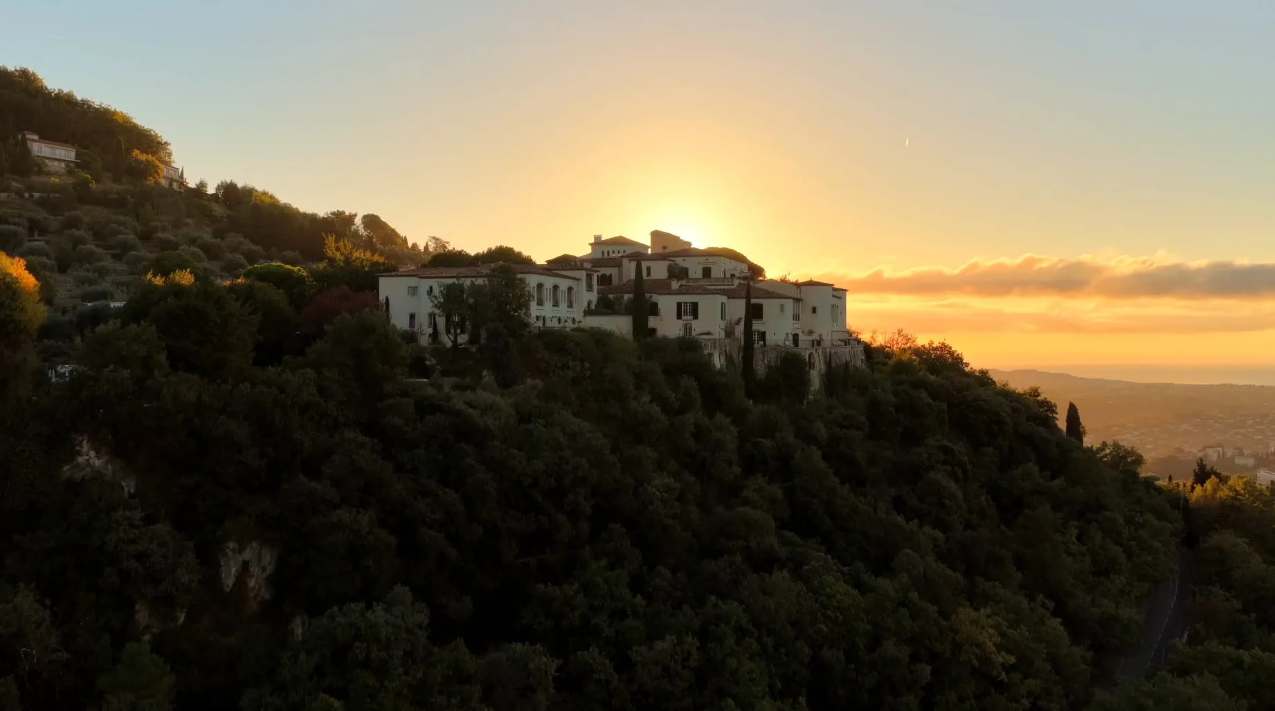 The stone facade of Chateau Saint-Martin & Spa rising above terraced olive groves on a hillside near Vence, with the Mediterranean coastline visible in the distance
