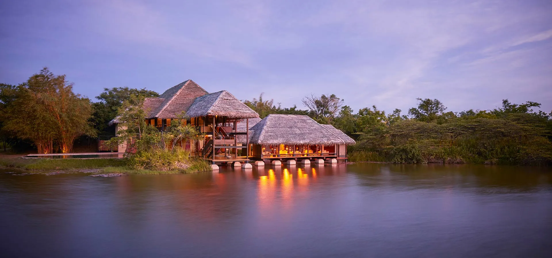 Thatched water dwellings on stilts above a still, reed-lined lake at Jetwing Vil Uyana, with Sigiriya rock visible in the distance