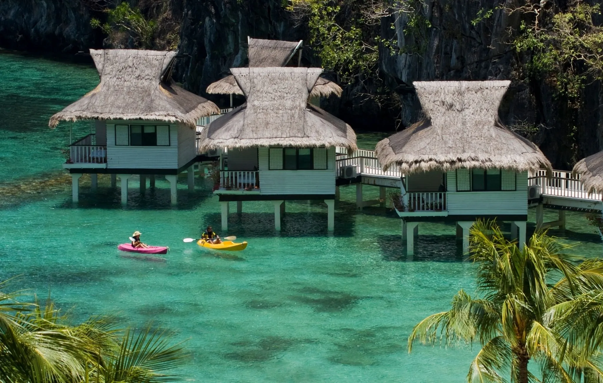 Thatched-roof water cottages on stilts at Miniloc Island Resort set against the limestone cliffs of Bacuit Bay